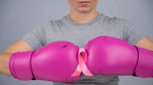 Caucasian woman in pink boxing gloves with a pink ribbon on her chest on a gray background. Fight against breast cancer