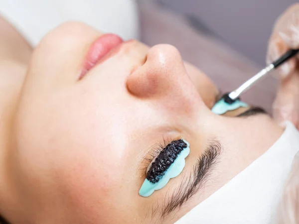 Close-up portrait of a woman on eyelash lamination procedure. The master applies tint to the eyelashes