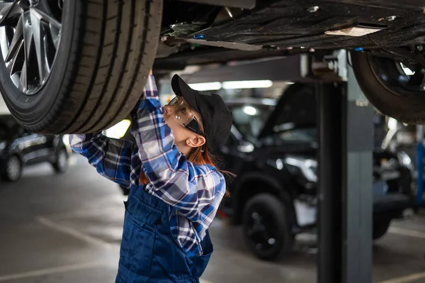 A female mechanic inspects a lifted car. A girl at a mans work