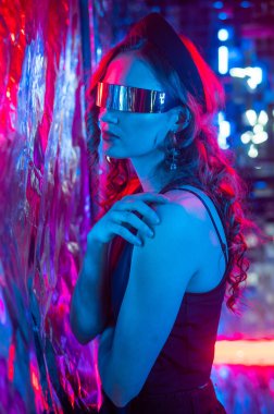 Beyaz tenli, güneş gözlüklü, neon ışıklı kadının folyo duvarına yakın plan portresi.