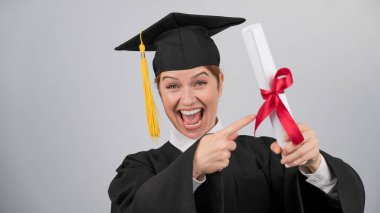 Mezuniyet elbisesi giymiş mutlu bir kadın diplomasını işaret ediyor.