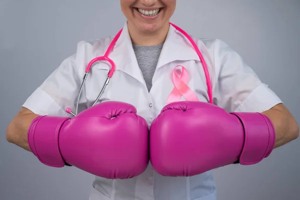 Smiling female doctor in pink boxing gloves with a pink ribbon on a gray background. Fight against breast cancer