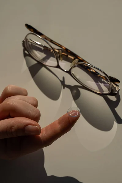 Close-up of a contact lens on a female index finger against the background of glasses on a white table