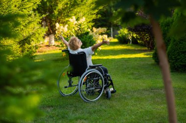 Rear view of an elderly woman spread her arms to the sides while sitting in a wheelchair on a walk outdoors