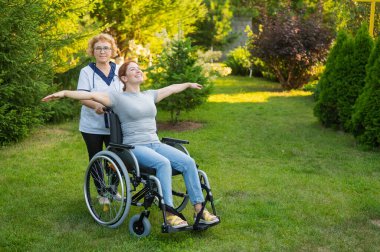 An elderly nurse walks with a middle-aged woman in a wheelchair through the park. The girl spread her arms outstretched like wings