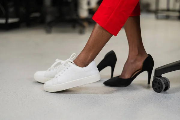 African woman changing from high heels to sneakers in the office. Tired legs