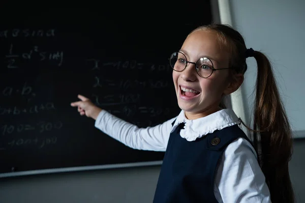 Portrait of a caucasian girl in glasses in the classroom. The schoolgirl writes the formula with chalk on the blackboard and looks into the camera