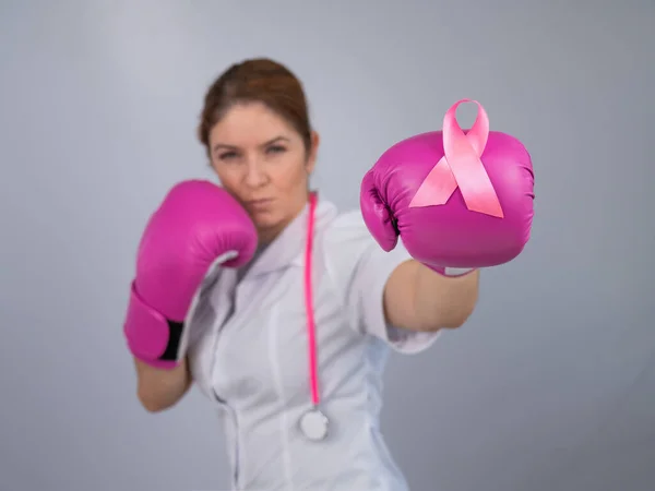 Female doctor in pink boxing gloves with a pink ribbon on a gray background. Fight against breast cancer