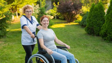An elderly female nurse walks with a middle-aged woman in a wheelchair in the park