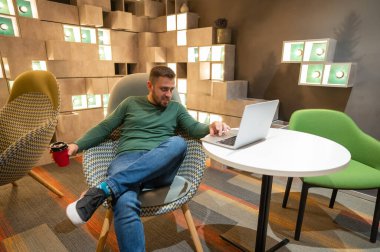 Caucasian bearded man sits in a comfortable chair drinks coffee and works on a laptop. Freelancer