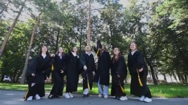 Classmates in graduation gowns throwing hats outdoors. Slow motion