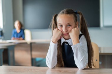 Küçük beyaz kız okuldaki dersten sıkıldı. Liseli kız masasında oturuyor ve öğretmen arka planda oturuyor.