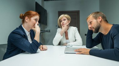 Sarışın, kızıl saçlı bir kadın ve ofiste takım elbiseli sakallı bir adam. Meslektaşlar konferans odasındaki bir masaya oturur ve kafalarını düşünceye daldırırlar.