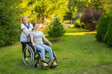 An elderly female nurse walks with a middle-aged woman in a wheelchair in the park