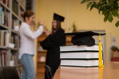 Kütüphanedeki bir yığın kitabın mezuniyet şapkasının yakın çekimi. Öğretmen ve genç bayan arka planda mezun oldular.