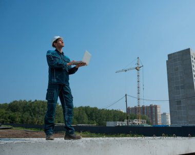 Kasklı bir erkek müteahhit elinde dizüstü bilgisayarla inşaat alanının arka planında duruyor.