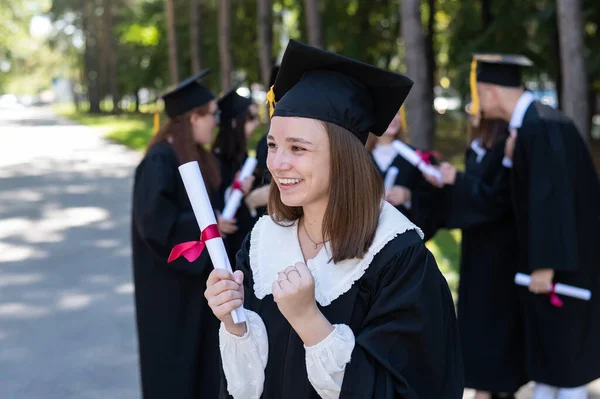 Group of happy students in graduation gowns outdoors. A young girl is happy to receive her diploma