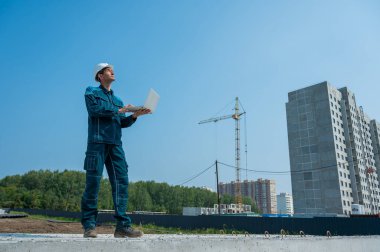 Kasklı bir erkek müteahhit elinde dizüstü bilgisayarla inşaat alanının arka planında duruyor.