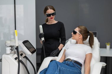 Gözlüklü bir kanepede bir kadın fotorojenizasyon ameliyatı oluyor. Kozmetikçinin hastası var.