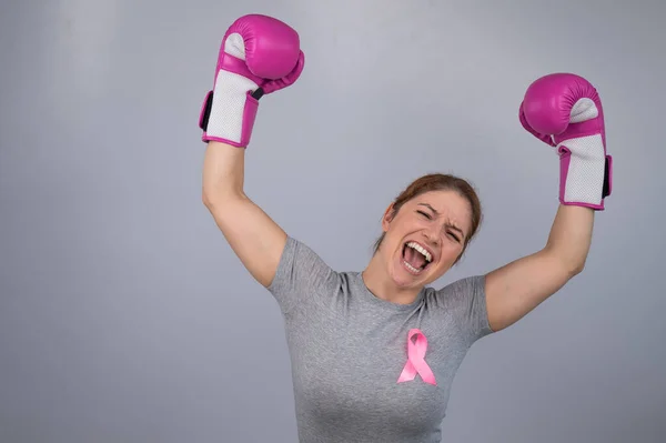 A woman with a pink ribbon on her chest raises her hands in pink boxing gloves on a gray background. Victory over breast cancer