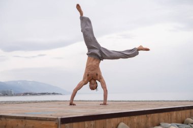 Üstsüz beyaz adam sahilde akrobatik tekerlek yapıyor.