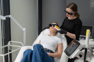 Gözlüklü genç beyaz kadın, fotojenizasyon prosedürü uyguluyor. Doktor kozmetik uzmanı ve kadın bir hasta.