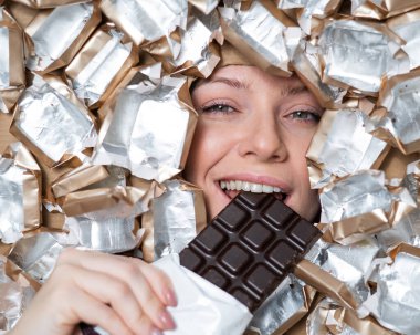 The face of a Caucasian woman surrounded by candy wrappers. The girl eats a bar of chocolate