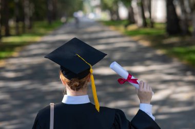 Rear view of a caucasian woman in a graduate gown holding a diploma outdoors