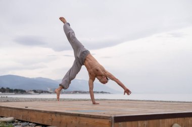 Üstsüz beyaz adam sahilde akrobatik tekerlek yapıyor.