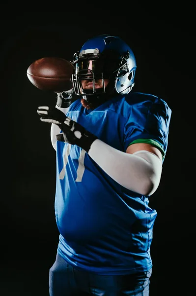 An Asian man with a red beard in a blue American football uniform throws a ball against a black background