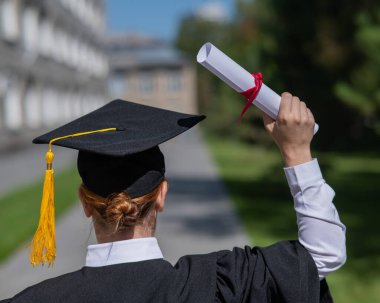 Mezuniyet elbisesi giymiş, elinde diplomasıyla üniversitenin yanında duran beyaz bir kadının arka görünüşü.