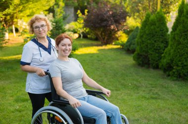 An elderly female nurse walks with a middle-aged woman in a wheelchair in the park
