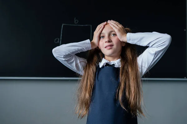 Caucasian girl stands at the blackboard holding her head with her hands. The schoolgirl did not prepare for the lesson