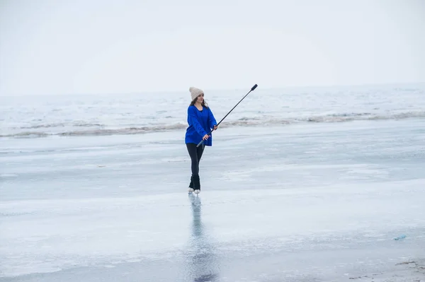 A caucasian woman is skating on a frozen lake holding a selfie stick in her hands. The figure skater films her skating