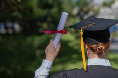 Mezuniyet elbisesi giymiş, elinde diplomasıyla üniversitenin yanında duran beyaz bir kadının arka görünüşü.