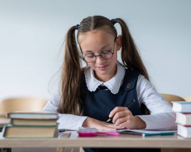 İki at kuyruklu beyaz bir kız masasında oturuyor ve bir çalışma kitabına yazıyor.