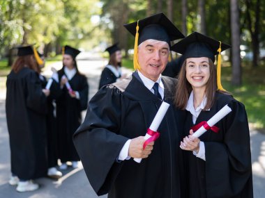 Bir grup mezun açık havada bornozla geziyor. Yaşlı bir adam ve genç bir kadın mezuniyetleri için birbirlerini kutluyorlar.