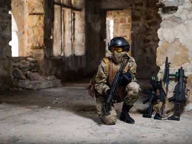 Askeri üniformalı bir kadın terk edilmiş bir binada oturuyor ve elinde ateşli bir silah tutuyor.