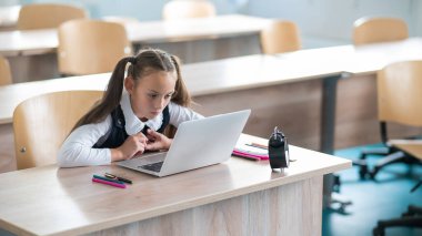 Beyaz kız okulda bir masada oturuyor ve bir dizüstü bilgisayarda okuyor. Masadaki çalar saat.