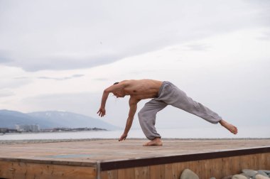 Üstsüz beyaz adam sahilde akrobatik tekerlek yapıyor.