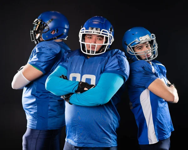 Portrait of three men in blue American football uniforms standing with their arms crossed over their chests on a black background