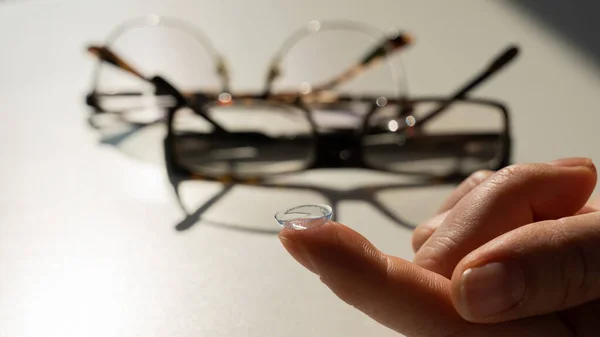 Close-up of a contact lens on a female index finger against the background of glasses on a white table