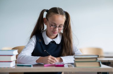 İki at kuyruklu beyaz bir kız masasında oturuyor ve bir çalışma kitabına yazıyor.