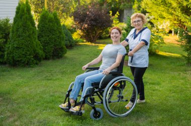 An elderly female nurse walks with a middle-aged woman in a wheelchair in the park