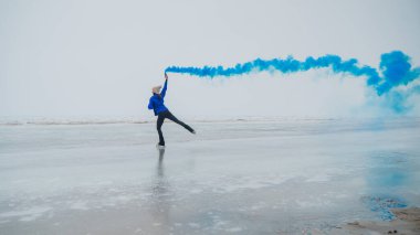 Beyaz bir kadın ellerinde renkli mavi bir dumanla donmuş bir gölde paten kayıyor.