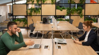 Kızıl saçlı beyaz kadın akıllı telefon kullanıyor ve sakallı adam da açık uzay ofisinde dizüstü bilgisayarın arkasında çalışıyor.