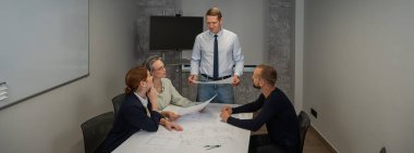 A caucasian man stands and holds a drawing, three colleagues sit at a table and listen to him