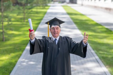 Mezuniyet bornozu giymiş yaşlı, kır saçlı bir adam kollarını yana açmış ve açık havada bir diploma tutuyor.