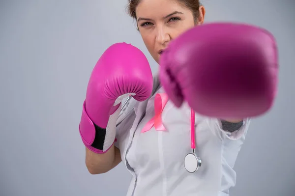 Serious female doctor in pink boxing gloves with a pink ribbon on a gray background. Fight against breast cancer