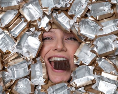 The face of a Caucasian woman surrounded by candy wrappers. The girl eats a bar of chocolate